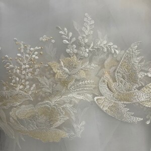 Op de afbeelding: Close-up van witte stof met geborduurd bloemen- en vogelontwerp. Het borduurwerk heeft cr&egrave;me- en goudkleurig garen, waardoor een delicaat en elegant patroon ontstaat. Het ontwerp omvat bloemen, bladeren en een gestileerde vogel.