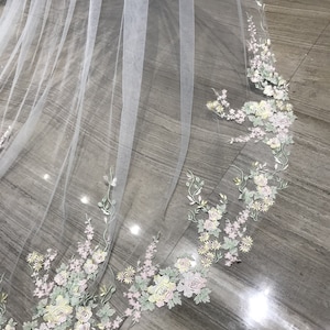 May include: A white tulle wedding veil with delicate floral embroidery in shades of pink, yellow, and green. The embroidery features a variety of flowers and leaves, creating a romantic and elegant look.