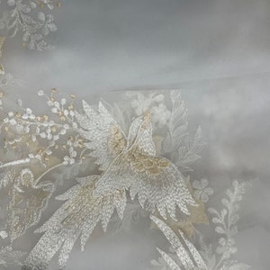 Op de afbeelding: Transparante witte stof met ingewikkelde borduursels. Het ontwerp toont een vogel en bloemmotieven in goud en zilver draad. De borduursels cre&euml;ren een textuureffect, ideaal voor bruids- of formele kleding.