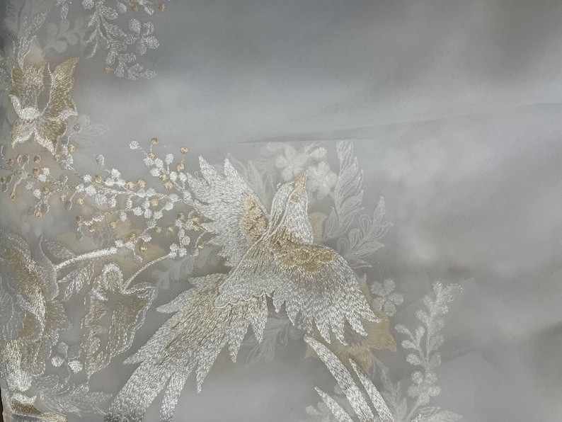 Op de afbeelding: Transparante witte stof met ingewikkelde borduursels. Het ontwerp toont een vogel en bloemmotieven in goud en zilver draad. De borduursels cre&euml;ren een textuureffect, ideaal voor bruids- of formele kleding.
