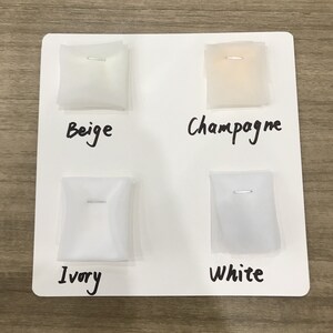 Op de afbeelding: Een witte kaart toont vier stofstalen in Beige, Champagne, Ivoor en Wit. Elk staal is vastgezet met een zilveren speld. De kleuren zijn beschreven met een handgeschreven zwarte marker.