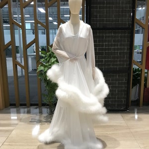 Puede incluir: Una bata blanca de gasa con una falda larga y fluida y mangas largas. La bata est&aacute; adornada con una gruesa banda de plumas blancas alrededor del dobladillo y los pu&ntilde;os.