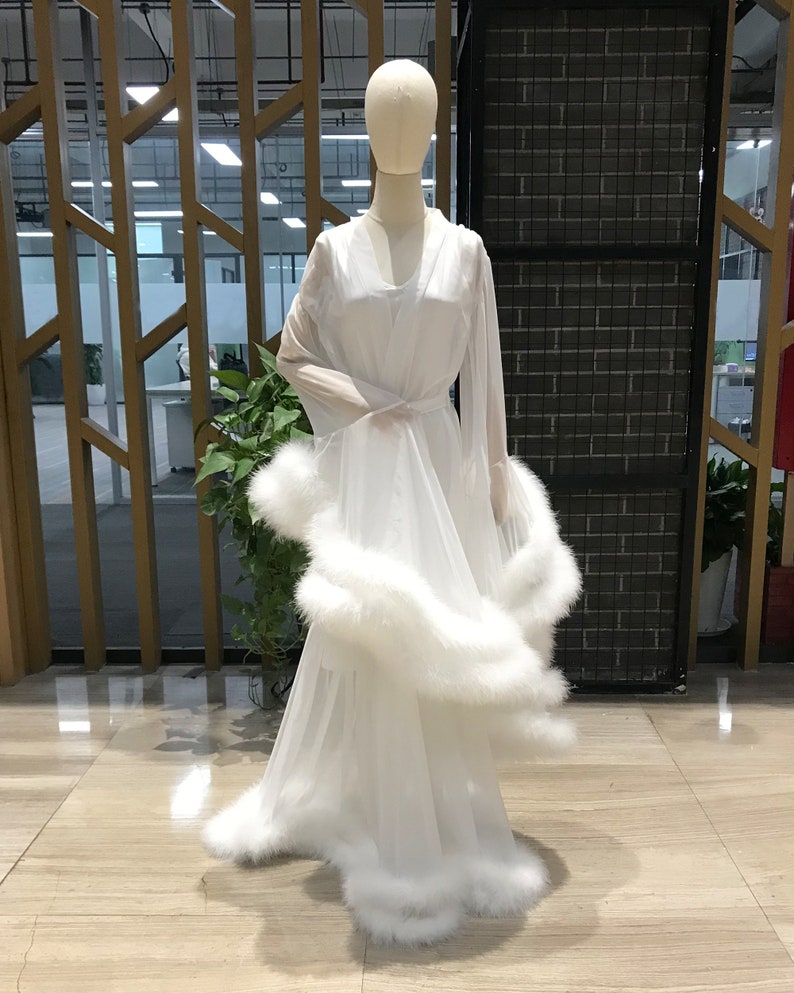 Puede incluir: Una bata blanca de gasa con una falda larga y fluida y mangas largas. La bata est&aacute; adornada con una gruesa banda de plumas blancas alrededor del dobladillo y los pu&ntilde;os.