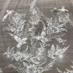 Op de afbeelding: Een witte bruidssluier met ingewikkelde bloemen- en vogelborduurwerk. Het delicate kanten ontwerp heeft gedetailleerde bloemen en vogels, wat een elegante en romantische esthetiek cre&euml;ert. De tekst "Ross Jack Co" is zichtbaar.