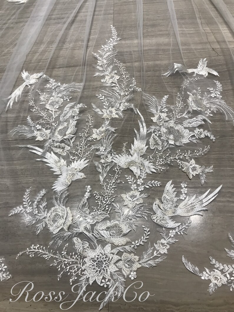 Op de afbeelding: Een witte bruidssluier met ingewikkelde bloemen- en vogelborduurwerk. Het delicate kanten ontwerp heeft gedetailleerde bloemen en vogels, wat een elegante en romantische esthetiek cre&euml;ert. De tekst "Ross Jack Co" is zichtbaar.