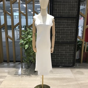 May include: A white satin slip dress with a V-neckline and adjustable straps. The dress is displayed on a mannequin on a gold stand.