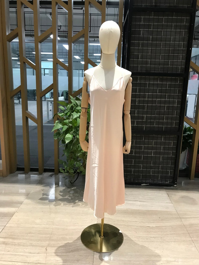 Puede incluir: Un vestido de sat&eacute;n rosa claro con tirantes finos. El vestido est&aacute; en un maniqu&iacute; y tiene un escote en V.