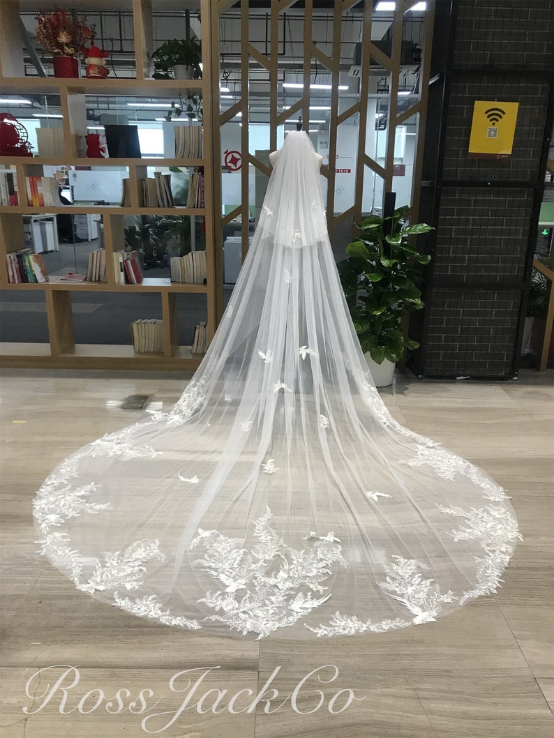 Op de afbeelding: Een lange, witte bruidssluier met delicate bloemen en vogelvormige kanten applicaties. De sluier wordt getoond op een mannequin, die het vloeiende ontwerp en de ingewikkelde details laat zien. De tekst "Ross Jack Co" is zichtbaar.