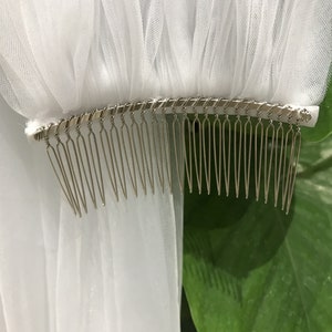 Op de afbeelding: Een witte bruidssluier met een metalen kam. De sluier is gemaakt van een transparante, vloeiende stof, verzameld aan de bovenkant en bevestigd aan een zilverkleurige metalen kam. De kam heeft veel dunne, gelijkmatig verdeelde tanden.
