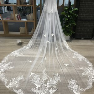 Op de afbeelding: Een lange, witte bruidssluier met ingewikkelde bloemen- en vogelachtige kanten details langs de randen. De sluier wordt op een mannequin getoond, waarbij de lengte en het delicate ontwerp worden benadrukt. De tekst "Ross Jack Co" is zichtbaar.