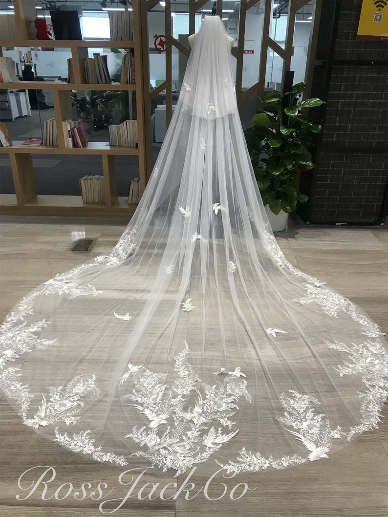 Op de afbeelding: Een lange, witte bruidssluier met ingewikkelde bloemen- en vogelachtige kanten details langs de randen. De sluier wordt op een mannequin getoond, waarbij de lengte en het delicate ontwerp worden benadrukt. De tekst "Ross Jack Co" is zichtbaar.