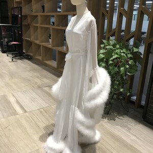 May include: A white sheer robe with a long sleeve and a white feather trim. The robe has a sash at the waist and a V-neckline.