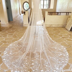 May include: A long, floor-length white bridal veil draped over a mannequin, featuring intricate floral lace embroidery along the hem and scattered throughout the tulle fabric. The text Ross Jack Co is printed in the center of the veil fabric.