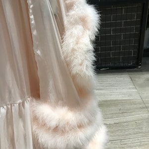 Puede incluir: Una bata blanca de gasa con un ribete de plumas blancas y esponjosas alrededor de los bordes. La bata tiene un dobladillo con volantes.