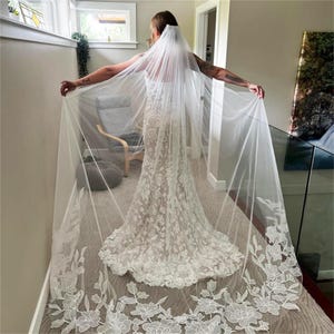 May include: A white wedding dress with a floral lace pattern and a long, sheer veil. The veil features a delicate floral lace trim along the edges. The dress has a fitted silhouette.