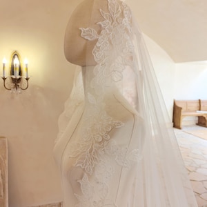 Puede incluir: Un velo de novia blanco con intrincados detalles de encaje floral está drapeado sobre un maniquí. El velo es transparente, permitiendo el paso de la luz, y el encaje se concentra a lo largo de los bordes y la parte superior. El maniquí está en una habitación luminosa.