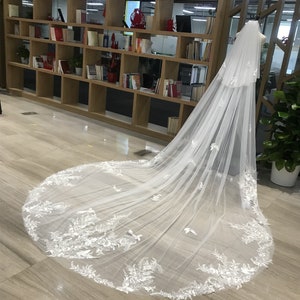 Op de afbeelding: Een lange, witte bruidssluier met ingewikkelde bloemen- en vlinderborduursels. De sluier wordt getoond op een mannequin, die het delicate ontwerp en de vloeiende stof laat zien. De sleep van de sluier strekt zich uit over de vloer en benadrukt de gedetailleerde kanten afwerking.