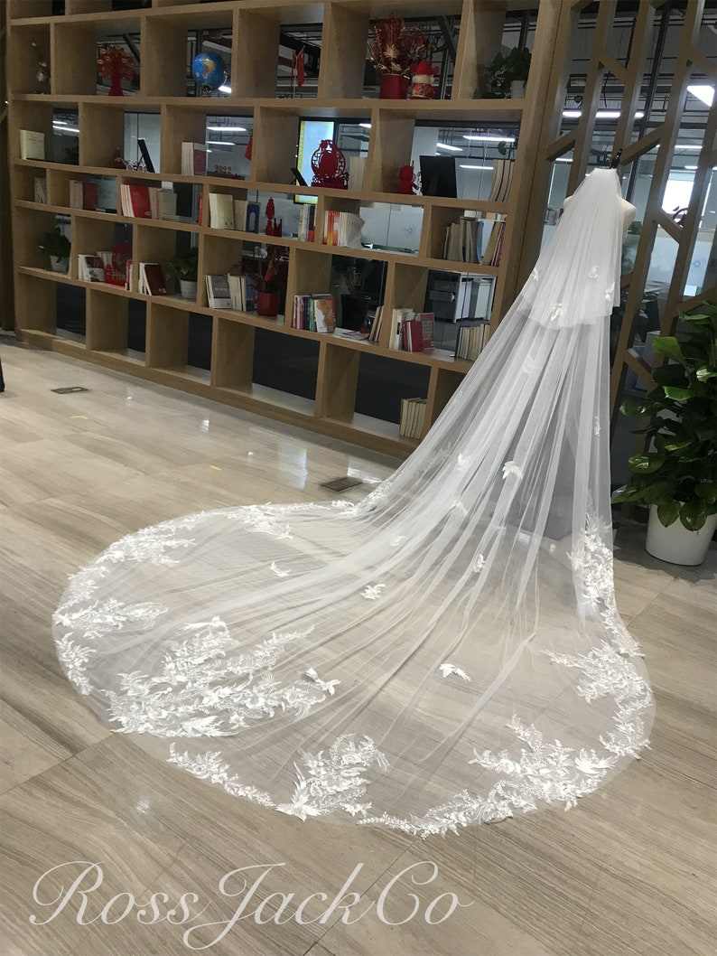 Op de afbeelding: Een lange, witte bruidssluier met ingewikkelde bloemen- en vlinderborduursels. De sluier wordt getoond op een mannequin, die het delicate ontwerp en de vloeiende stof laat zien. De sleep van de sluier strekt zich uit over de vloer en benadrukt de gedetailleerde kanten afwerking.