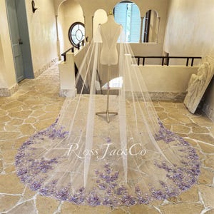 May include: A long, sheer white bridal veil with intricate lavender floral embroidery along the hem. The veil is displayed on a mannequin, showcasing its delicate design and the text "Ross Jack Co".