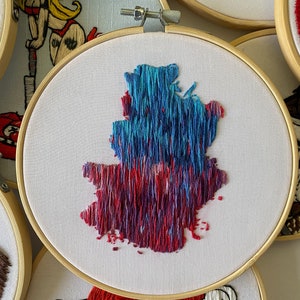 May include: Embroidered hoop art featuring a red and blue ombre design of the island of Ireland. The hoop is made of wood and has a white fabric background.
