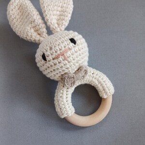 May include: A crocheted bunny rattle with a wooden ring. The bunny is cream coloured with black eyes and a brown bow tie.