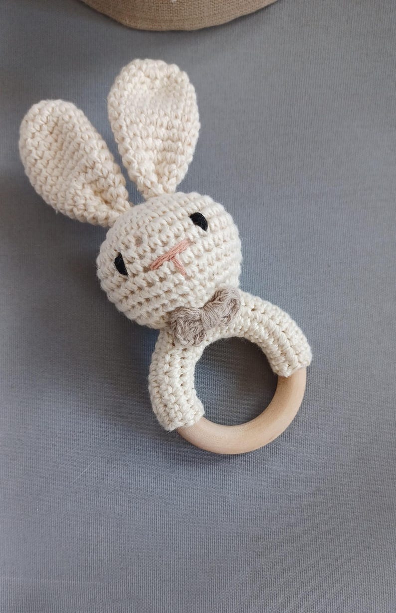 May include: A crocheted bunny rattle with a wooden ring. The bunny is cream coloured with black eyes and a brown bow tie.
