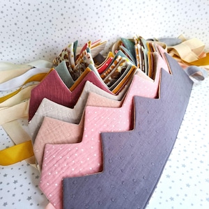 Könnte beinhalten: Eine Sammlung handgefertigter Stoffkronen in verschiedenen Farben, darunter Rosa, Grau und Burgunderrot. Jede Krone hat ein Zickzack-Design und ist mit Bändern in Gelb-, Creme- und Grautönen verziert. Die Kronen sind wahrscheinlich für Kinder oder Mottopartys gedacht.