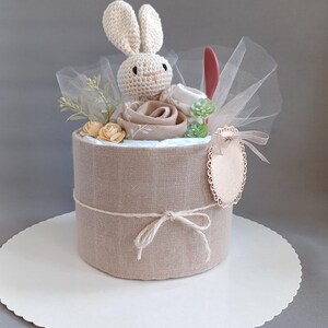 May include: A beige diaper cake with a crocheted bunny on top, surrounded by brown fabric roses and greenery. The cake is tied with twine and has a wooden tag with a scalloped edge.