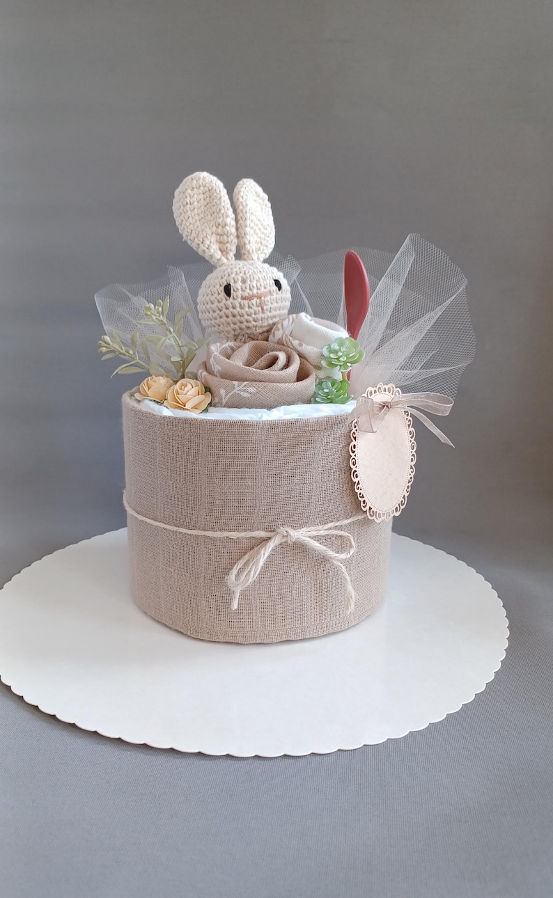 May include: A beige diaper cake with a crocheted bunny on top, surrounded by brown fabric roses and greenery. The cake is tied with twine and has a wooden tag with a scalloped edge.