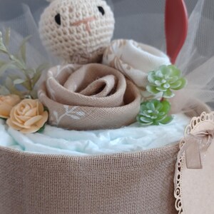 May include: A diaper cake decorated with a crocheted bunny, a brown fabric rose, and green succulents. The cake is wrapped in burlap and tied with twine. A small, brown, scalloped tag is attached to the side.