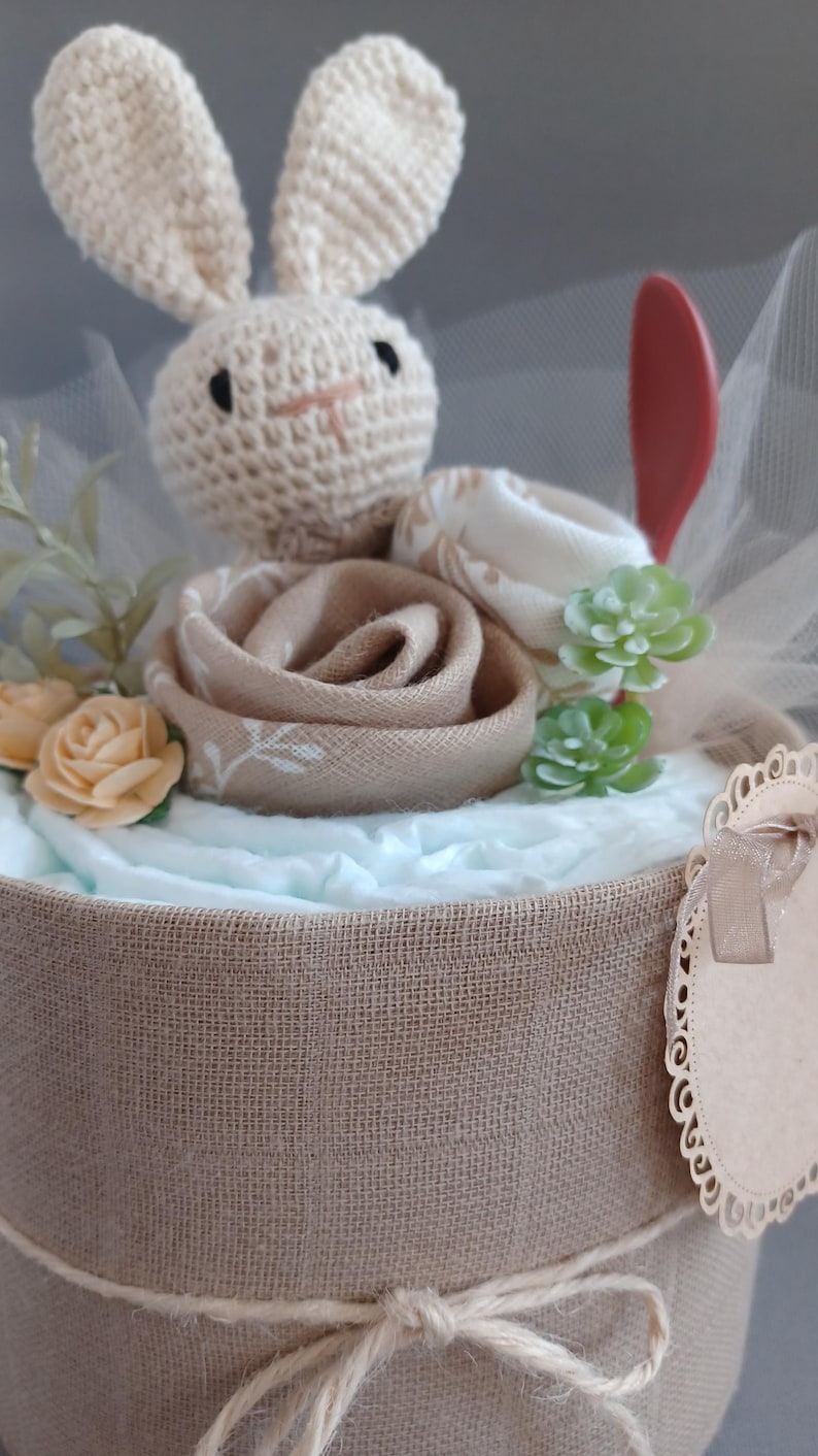 May include: A diaper cake decorated with a crocheted bunny, a brown fabric rose, and green succulents. The cake is wrapped in burlap and tied with twine. A small, brown, scalloped tag is attached to the side.