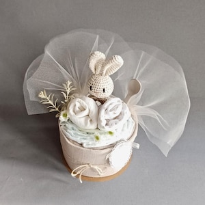 May include: A white crocheted bunny toy sits on top of a nappy cake wrapped in a beige fabric. The cake is decorated with white flowers and a white tulle bow.