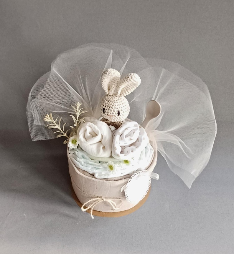 May include: A white crocheted bunny toy sits on top of a nappy cake wrapped in a beige fabric. The cake is decorated with white flowers and a white tulle bow.
