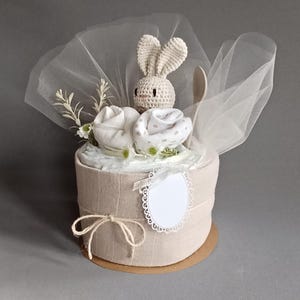 May include: A beige diaper cake decorated with white and beige fabric, a crocheted bunny toy, and a wooden spoon. The cake is wrapped in a sheer white fabric and tied with twine.