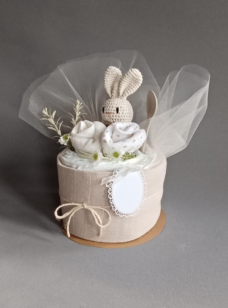 May include: A beige diaper cake decorated with white and beige fabric, a crocheted bunny toy, and a wooden spoon. The cake is wrapped in a sheer white fabric and tied with twine.