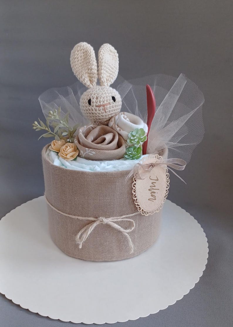 May include: A diaper cake decorated with a crocheted bunny, a wooden tag with the name "Julia", and a brown burlap base. The cake is decorated with yellow flowers and green succulents.