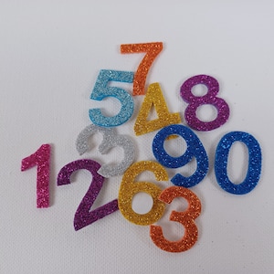 Può includere: Un set di numeri di schiuma colorati e scintillanti, tra cui 1, 2, 3, 4, 5, 6, 7, 8, 9 e 0. I numeri sono disposti in uno schema casuale su uno sfondo bianco.