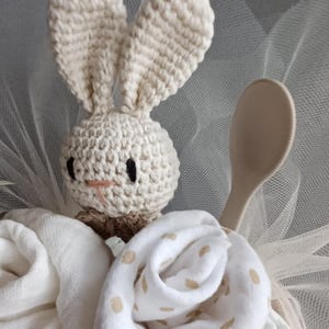 May include: A crocheted bunny toy with black eyes and a pink nose, sitting on top of a white and beige baby shower gift basket. The basket is filled with white and beige baby clothes and a wooden spoon.