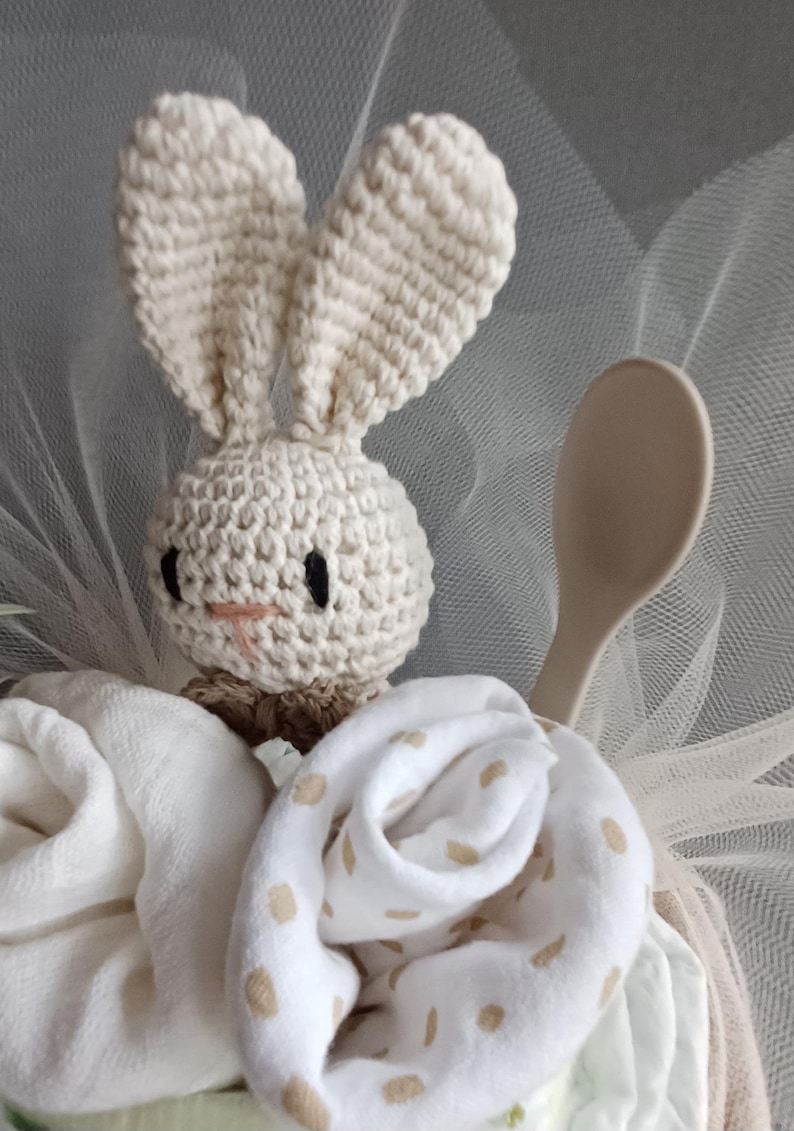May include: A crocheted bunny toy with black eyes and a pink nose, sitting on top of a white and beige baby shower gift basket. The basket is filled with white and beige baby clothes and a wooden spoon.