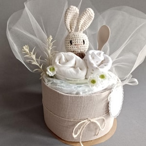 May include: A beige diaper cake with a crocheted bunny on top, surrounded by white and beige baby clothes, a wooden spoon, and white flowers. The cake is wrapped in a sheer white fabric and tied with a brown twine.