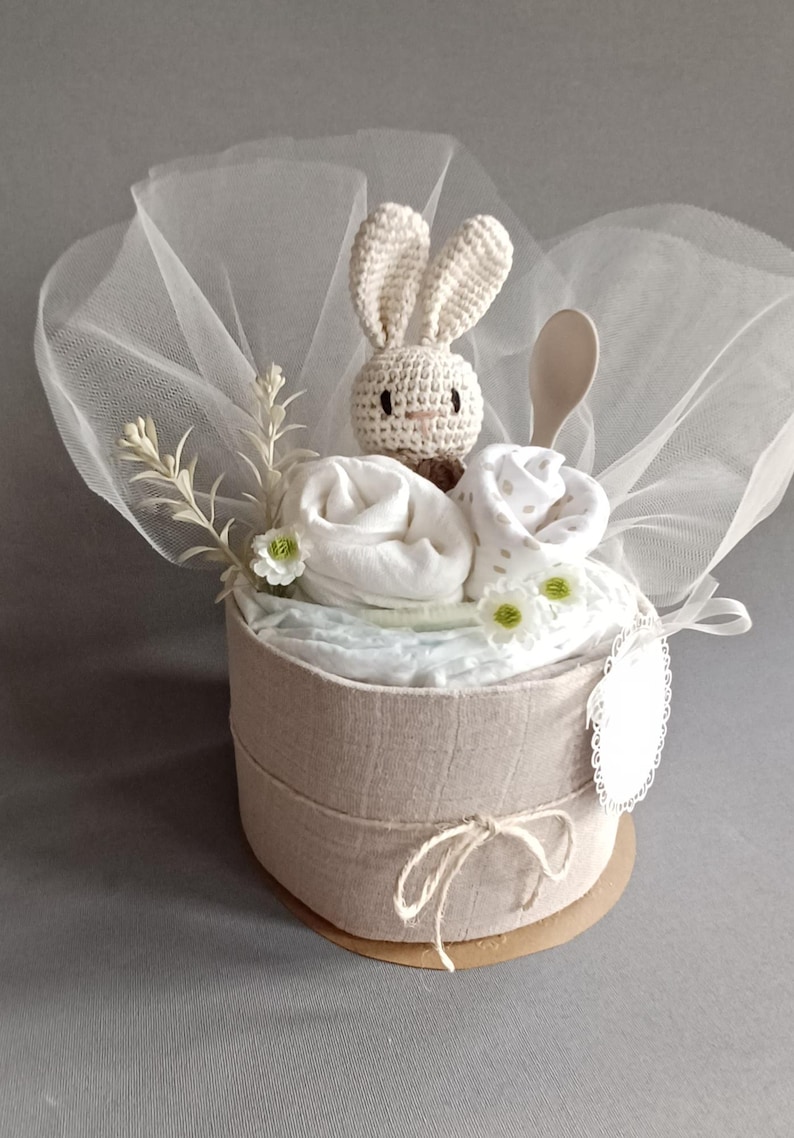 May include: A beige diaper cake with a crocheted bunny on top, surrounded by white and beige baby clothes, a wooden spoon, and white flowers. The cake is wrapped in a sheer white fabric and tied with a brown twine.