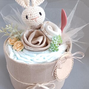 May include: A diaper cake decorated with a crocheted bunny, a brown fabric rose, a white fabric flower, and green succulents. The cake is wrapped in burlap and tied with twine. A small wooden tag with a scalloped edge is attached to the cake.
