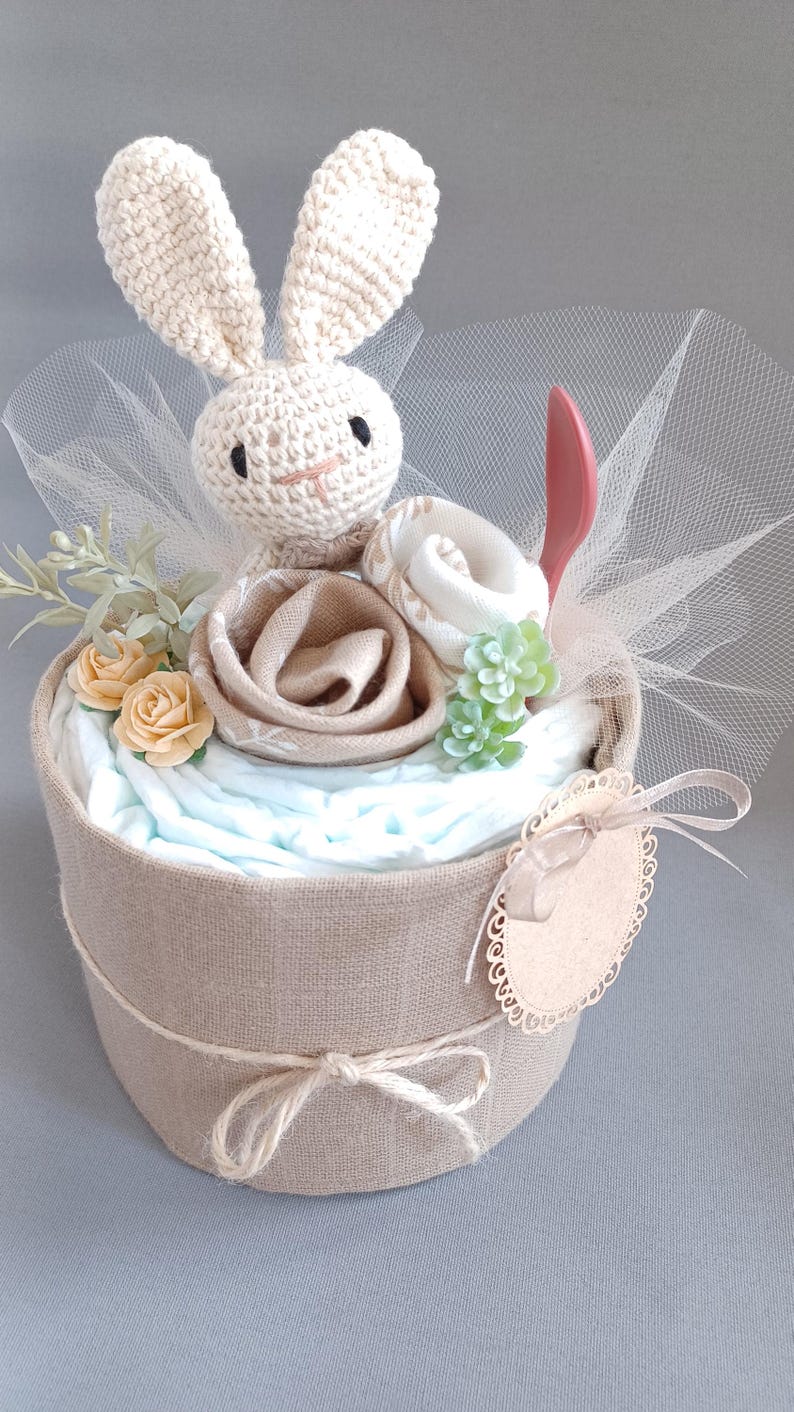 May include: A diaper cake decorated with a crocheted bunny, a brown fabric rose, a white fabric flower, and green succulents. The cake is wrapped in burlap and tied with twine. A small wooden tag with a scalloped edge is attached to the cake.