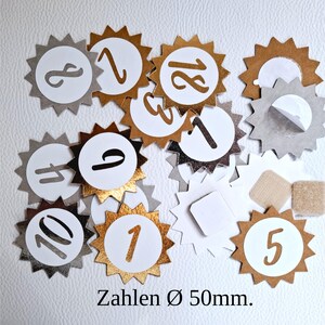 Könnte beinhalten: Eine Sammlung dekorativer Zahlenaufkleber. Die Aufkleber zeigen einen weißen Kreis mit einer Zahl in einem goldenen oder silbernen Sonnenstrahl-Design. Der Text "Zahlen Ø 50mm" ist unten zu sehen.