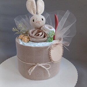 May include: A diaper cake decorated with a crocheted bunny, a small wooden sign, and a tulle bow. The cake is wrapped in burlap fabric and has a white base.