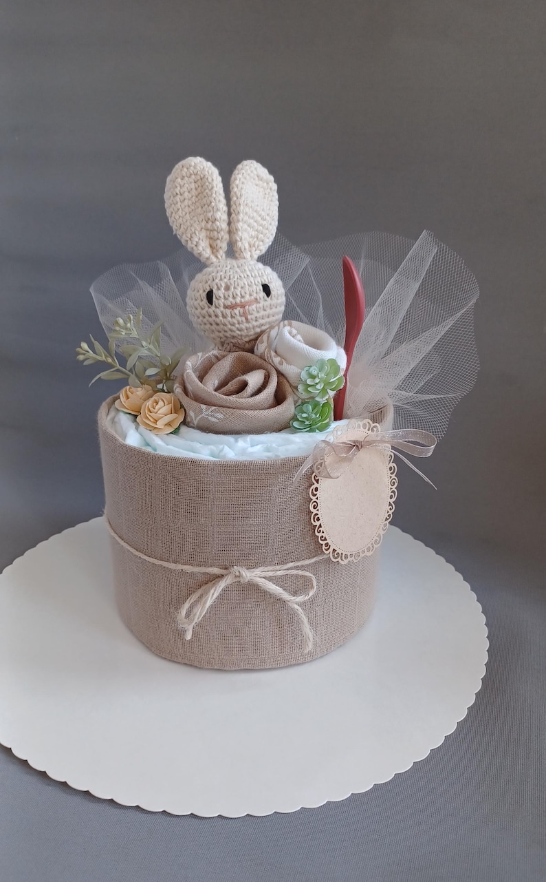May include: A diaper cake decorated with a crocheted bunny, a small wooden sign, and a tulle bow. The cake is wrapped in burlap fabric and has a white base.