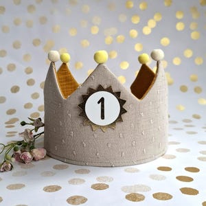 Könnte beinhalten: Eine beige Stoffkrone mit einem goldenen Stern und der Zahl 1. Die Krone hat ein gepunktetes Muster und ist mit gelbem Stoff verziert. Sie ist mit gelben und weißen Pompons verziert.