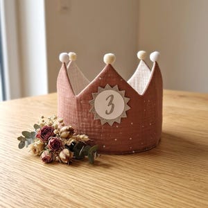 Pode incluir: Uma coroa de tecido castanho com bolinhas brancas e o número 3 prateado na frente. A coroa tem detalhes brancos e bege e é decorada com pequenos pompons brancos. Um pequeno buquê de flores secas está em frente à coroa.