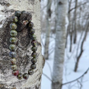 May include: A green, red, and brown beaded bracelet hangs from a birch tree in a snowy forest. The bracelet is made of natural stone beads.