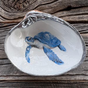 May include: A decorative seashell with a painted blue sea turtle inside. The shell is white with a silver trim and sits on a weathered wooden surface. The turtle has detailed blue markings.